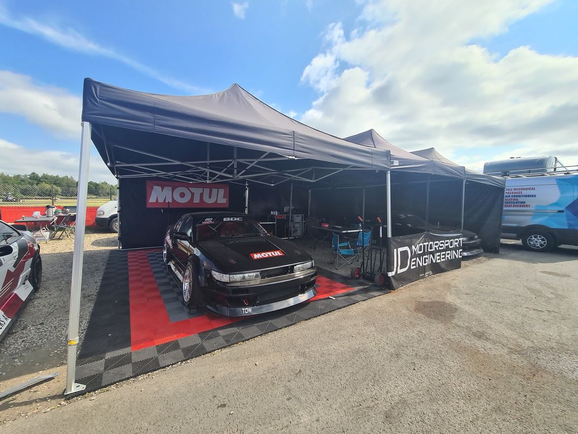 A black drift car sits beneath a gazebo canopy, on red and black swisstrax garage flooring